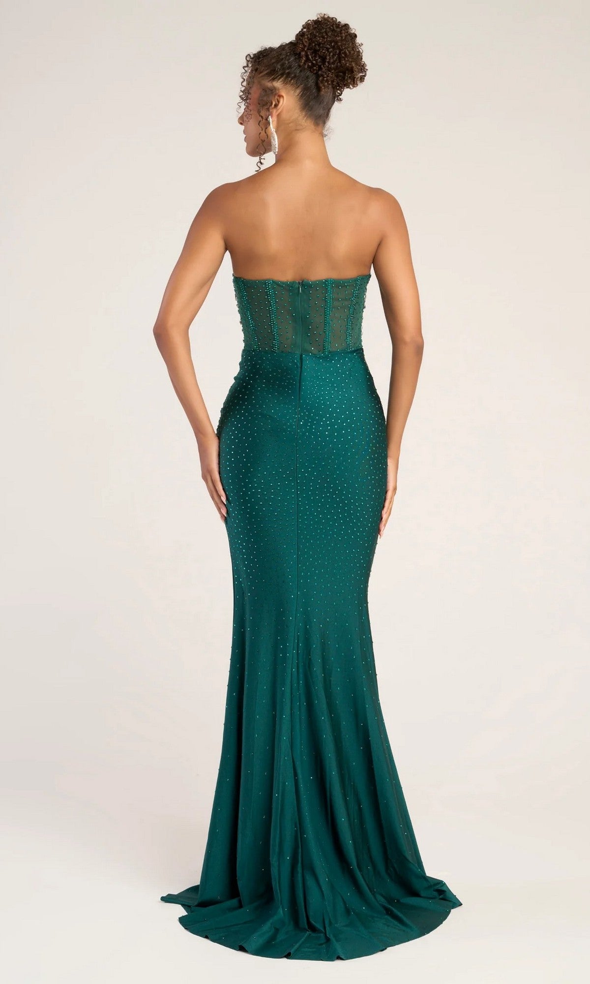A woman stands facing away, wearing the Portia and Scarlett XO012 long formal dress in green with a fitted silhouette, sparkling details, and a visible sheer corset bodice that highlights its elegant structure.